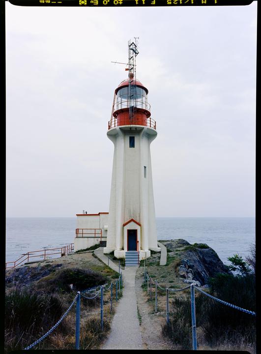 A white lighthouse with a red top stands on a rocky cliff overlooking the ocean under a cloudy sky.