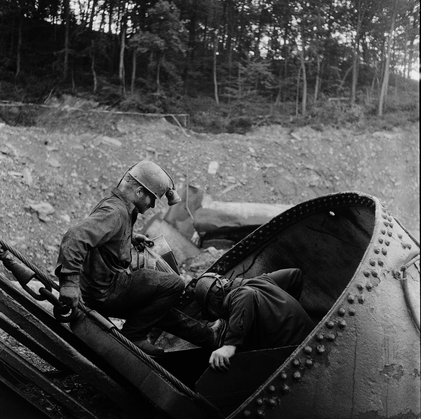 Two miners wearing hard hats and headlamps are working with heavy machinery in a wooded, rocky area.