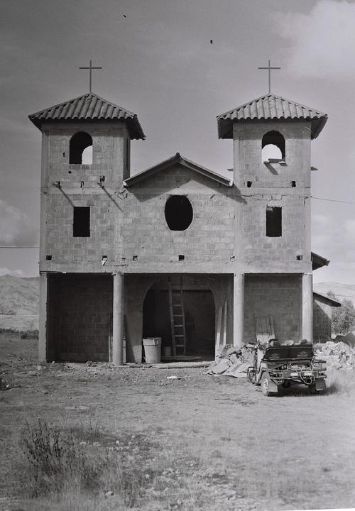 A two-towered church under construction stands in a field with a small vehicle parked in front.