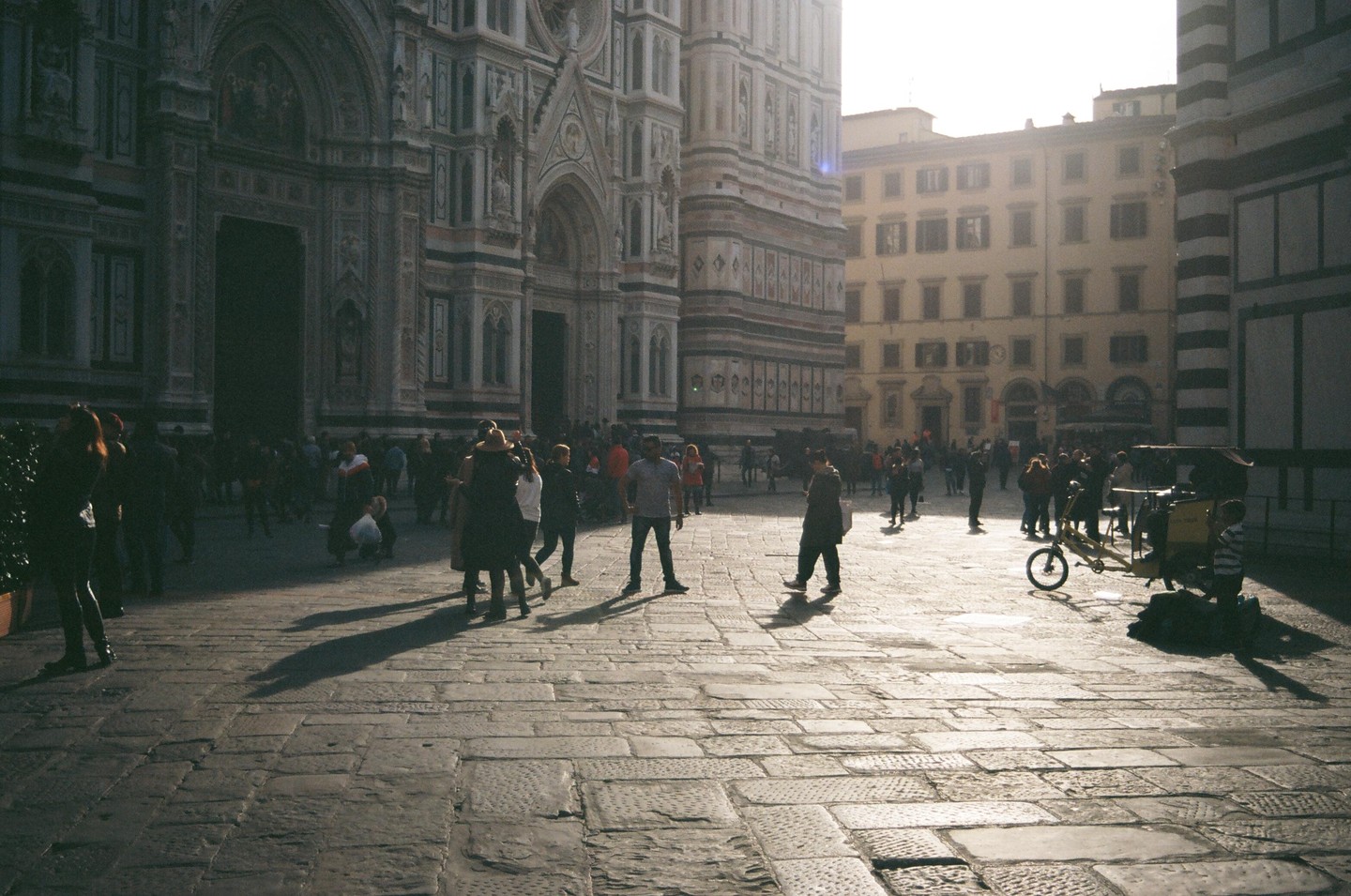 A crowd of people stands on a stone plaza in front of a large ornate building with another building in the background.