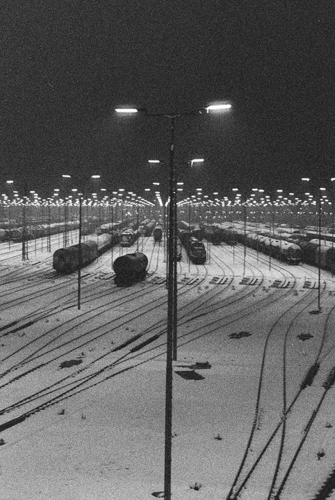 A train station is covered in snow at night, with many bright lights illuminating the tracks and rail cars.