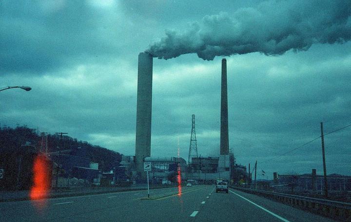 Smoke billows from two tall industrial smokestacks next to a highway under a cloudy sky.