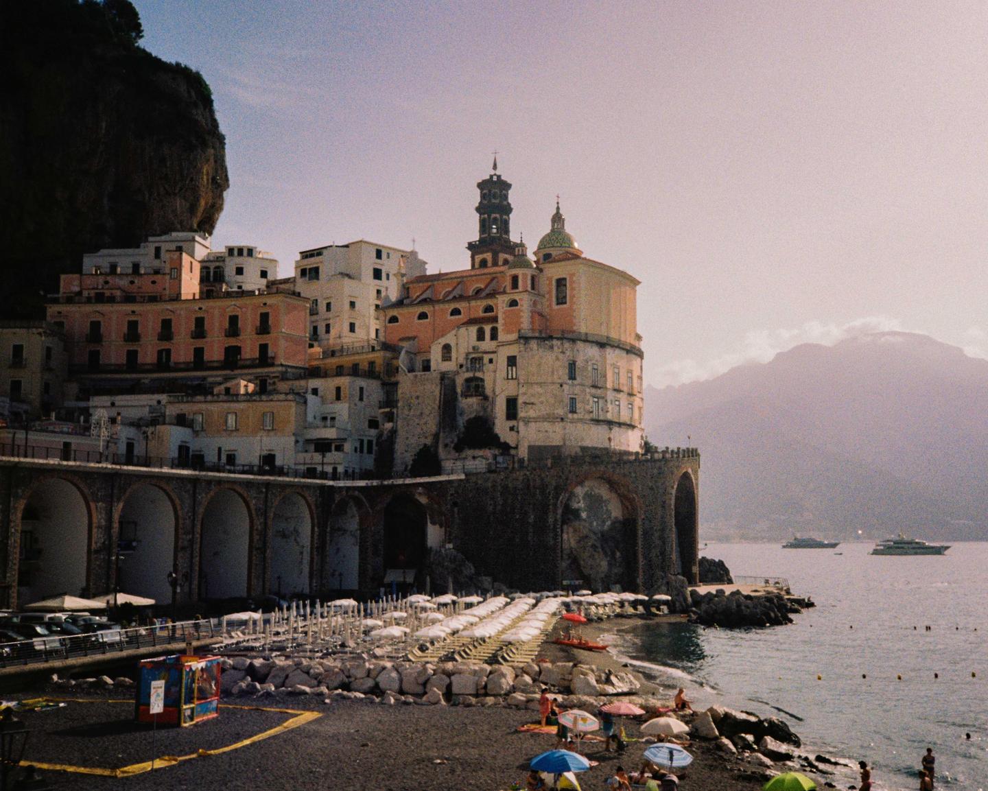 A coastal town with buildings built into a cliffside overlooks a beach with lounge chairs and umbrellas.