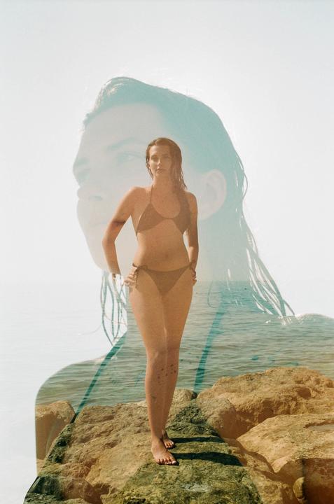 A woman in a bikini stands on rocks by the ocean with a faint double exposure of a woman's face in the background.