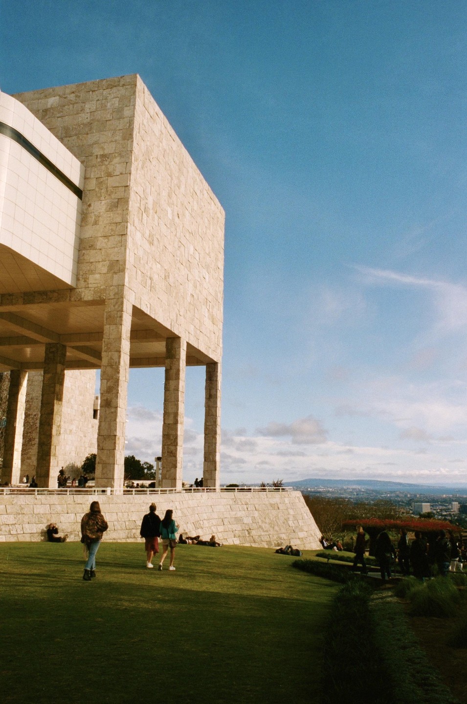 People relax on a grassy hill next to a large stone building with columns under a blue sky.