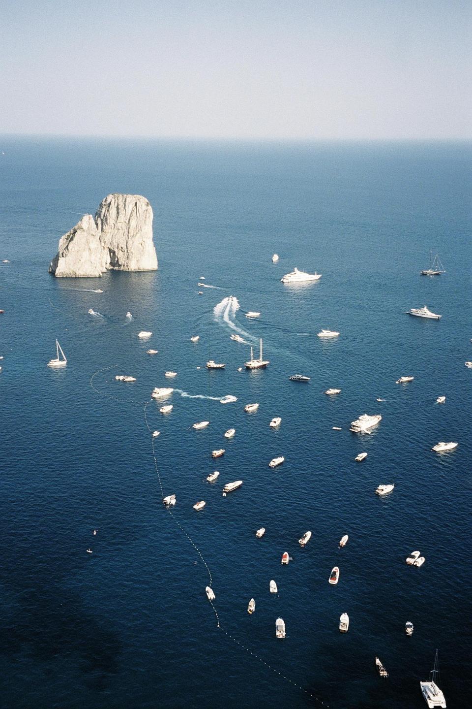 A large rock formation rises from the sea, surrounded by numerous boats and yachts.