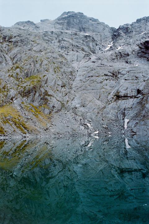 A mountain with patches of snow and moss is reflected in a still lake.