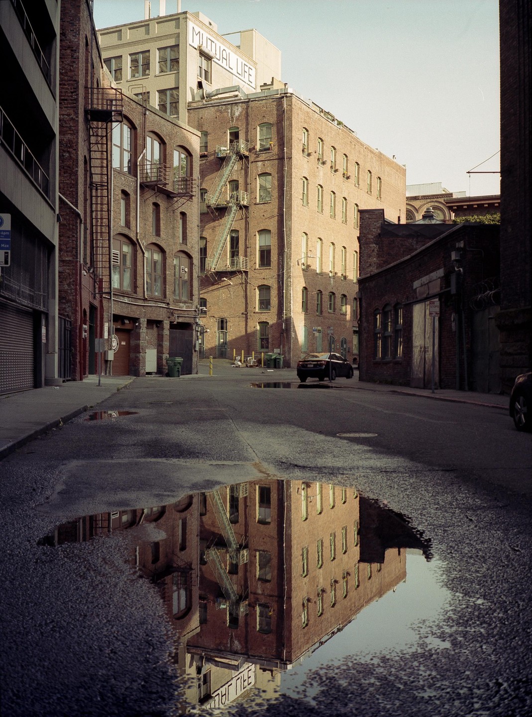 A street with brick buildings and fire escapes is reflected in a large puddle in the foreground.