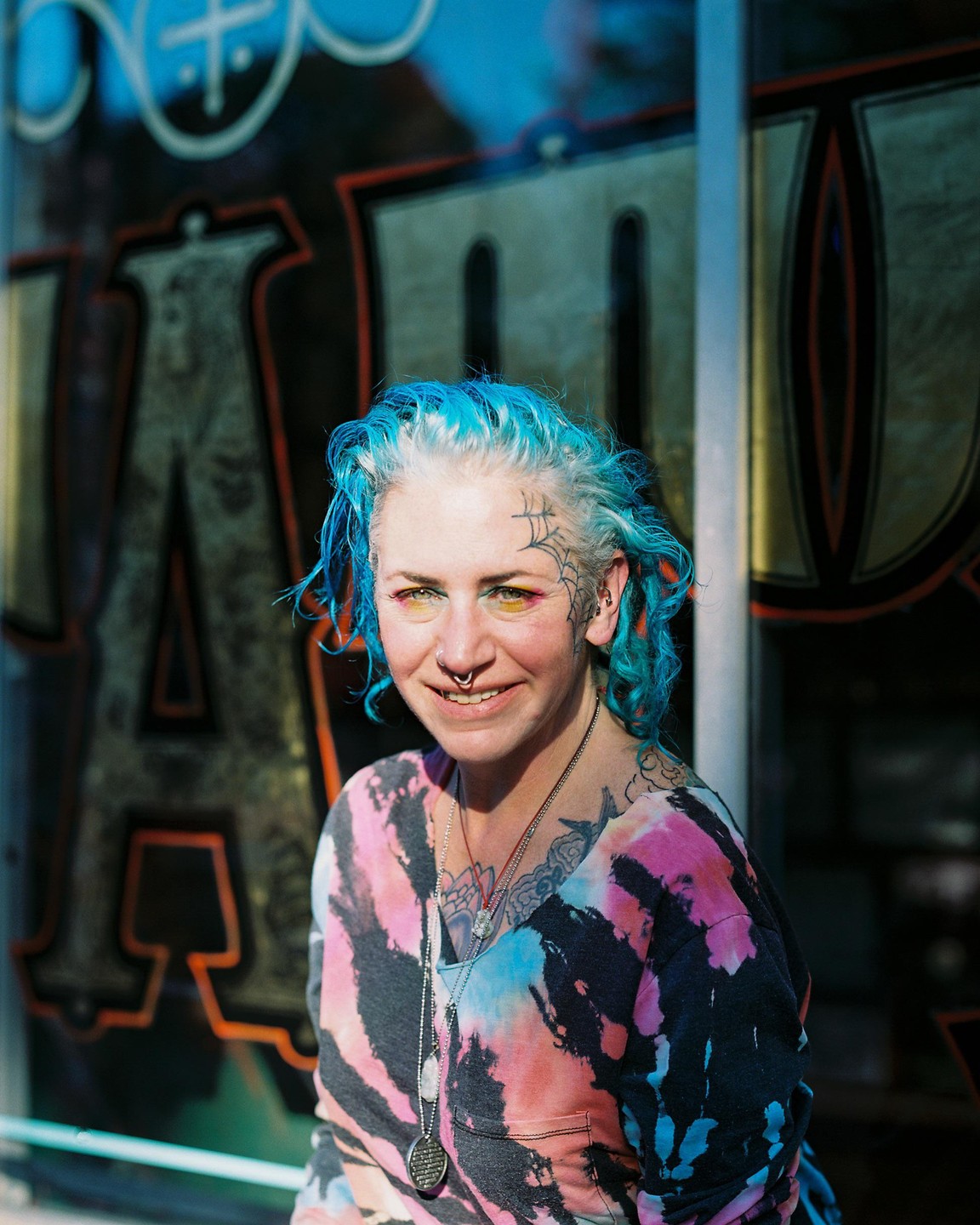 A woman with blue hair and a tie-dye shirt smiles at the camera.