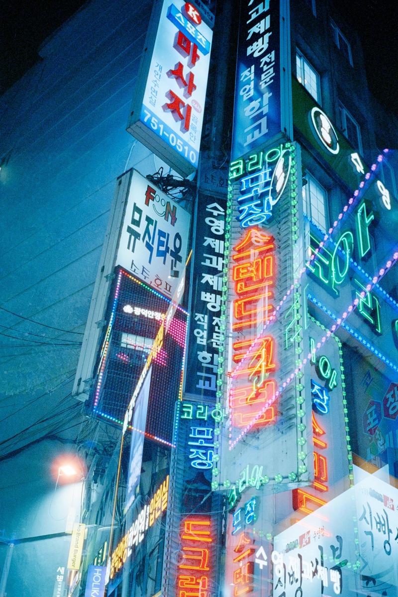 Multiple neon signs with Korean text are stacked vertically on the side of a building at night.