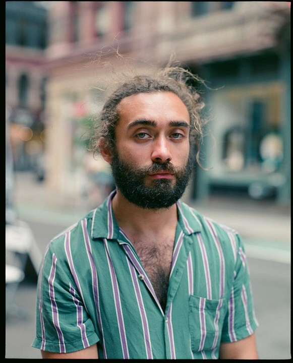 A man with a beard and curly hair wears a striped shirt and looks directly at the viewer on a city street.