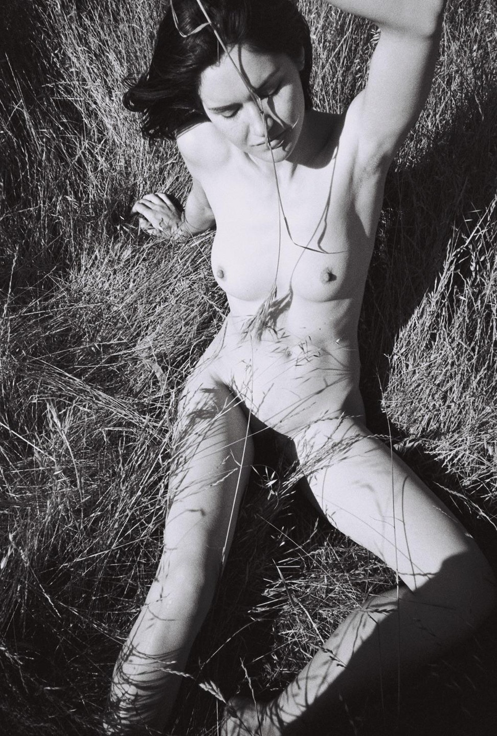 A nude woman is lying in tall grass with her eyes closed and her arm raised.