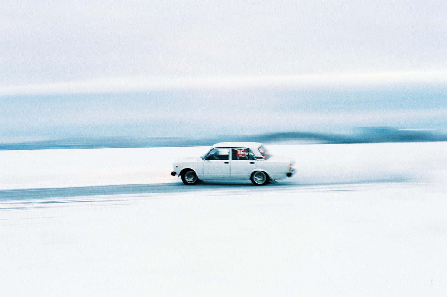 A white car with the number 16 on its side drives across a vast, snow-covered plain under a light blue sky.