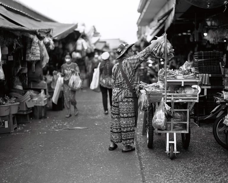 A woman wearing a hat and patterned pants is standing next to a cart filled with goods on a street.