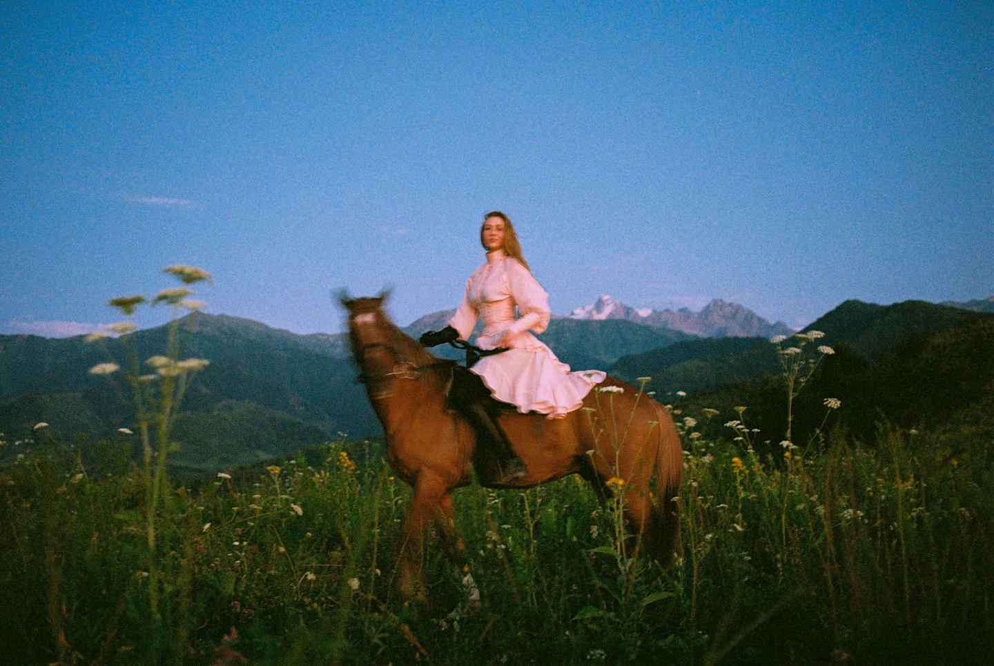 A woman in a pink dress rides a horse through a field of wildflowers with mountains in the background.