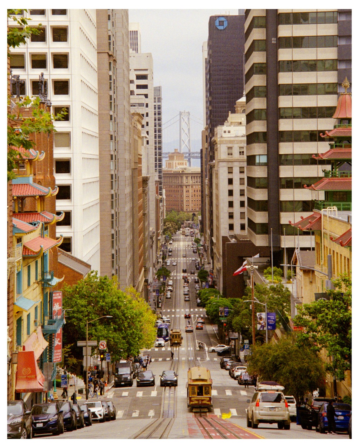 A cable car travels down a steep street lined with buildings and cars, with a bridge visible in the distance.