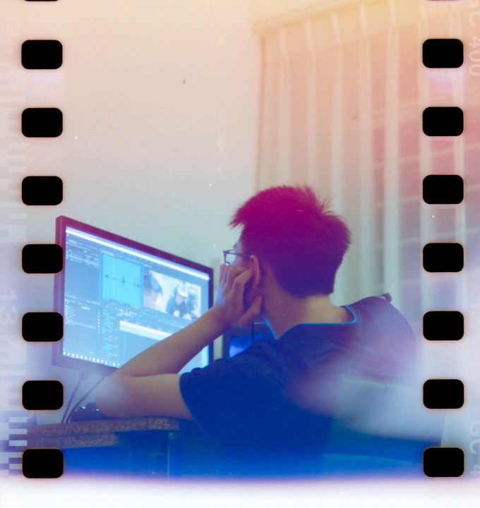 A man with spiky hair and glasses sits at a desk, looking at a computer monitor displaying video editing software.