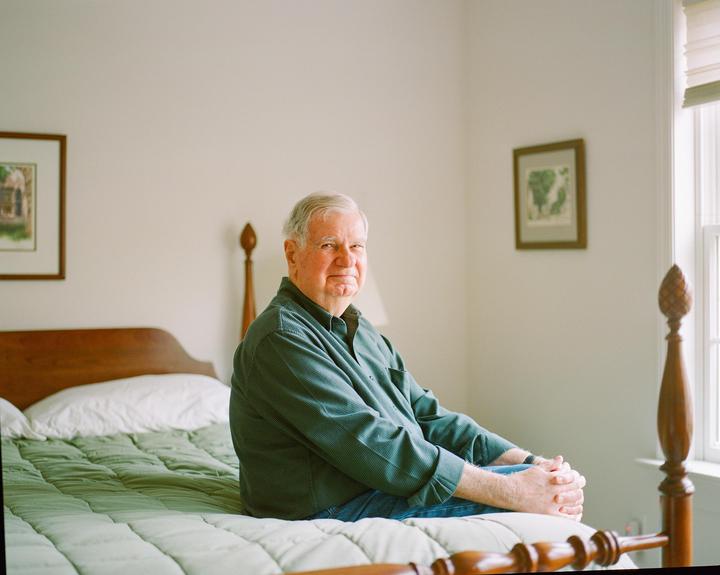 A man sits on a bed with his hands clasped in front of him.