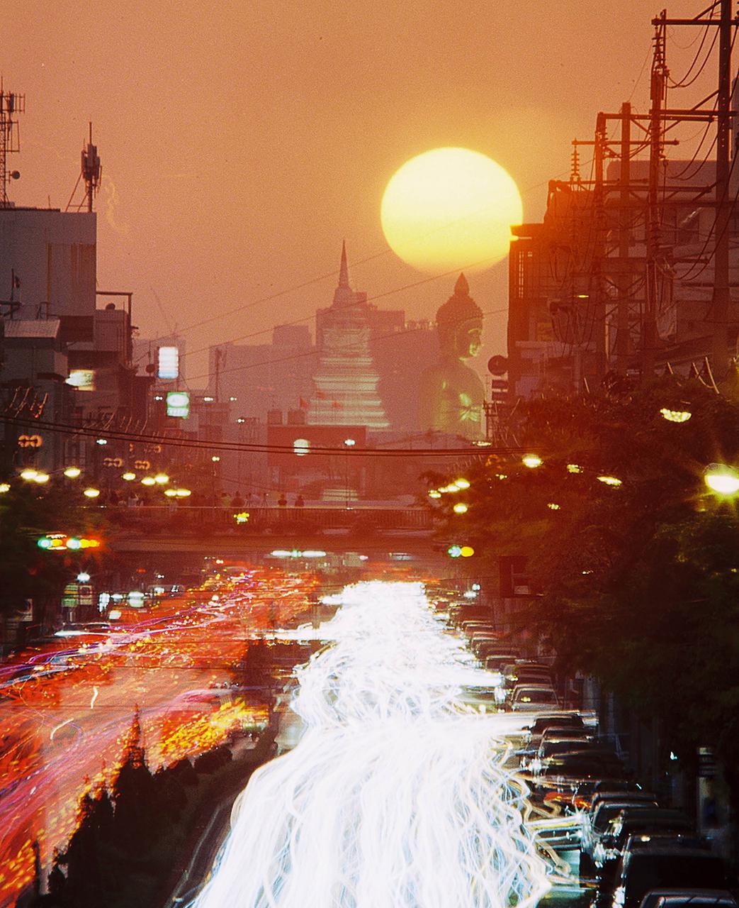 A large sun sets over a city street with light trails from moving vehicles and a large buddha statue in the background.
