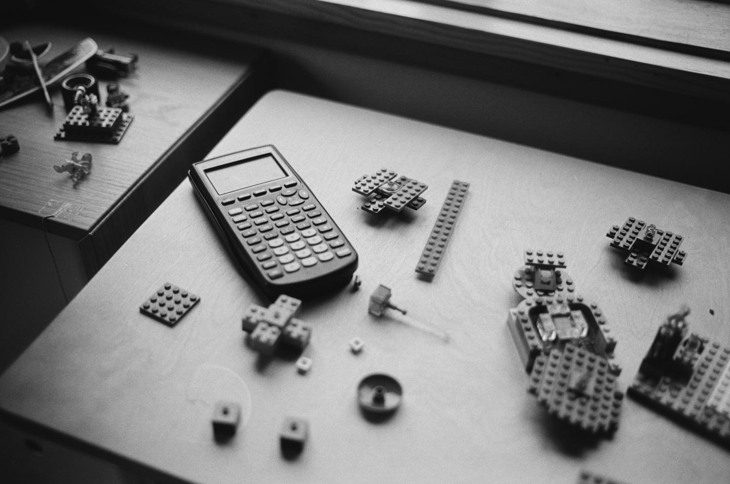 A calculator and various Lego pieces are scattered on a wooden surface next to a window.