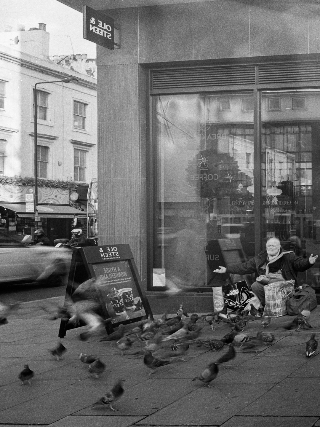 A man sits on the sidewalk feeding pigeons in front of a storefront.