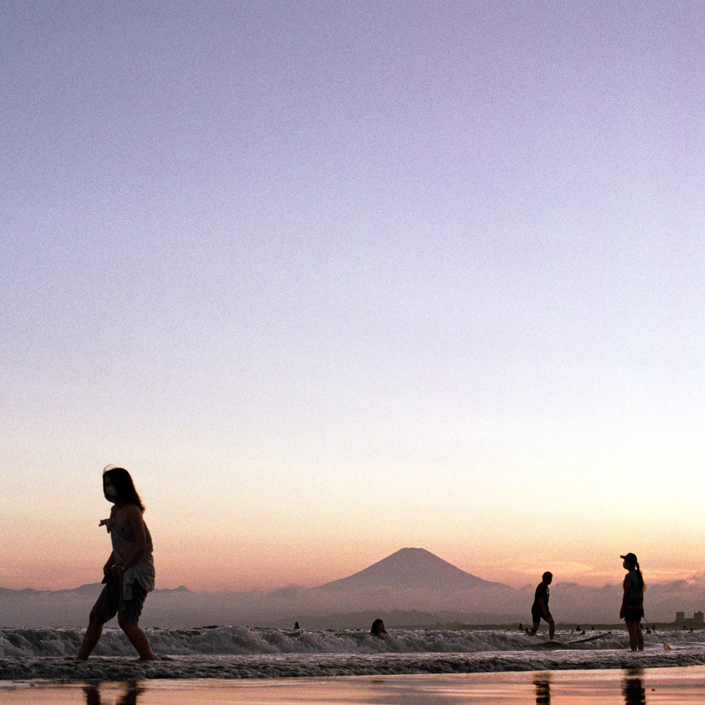 Silhouetted people stand in the ocean at sunset with a mountain in the distance.
