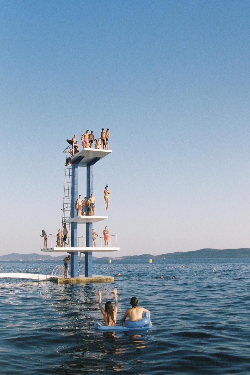 A group of people are on a tall diving platform in the middle of a body of water, with two people floating on inflatables in the foreground.
