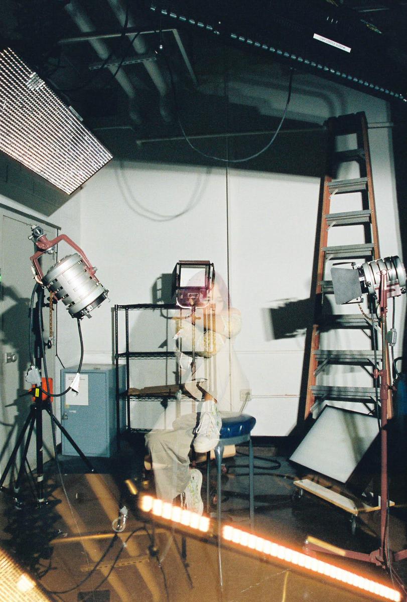 A person sits on a stool in a studio surrounded by various lighting equipment and a ladder.