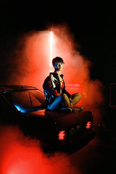 A woman sits on the hood of a car at night, illuminated by red and blue light.