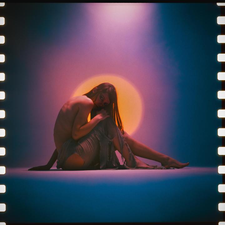 A woman sits on the floor with a bright orange circle behind her.