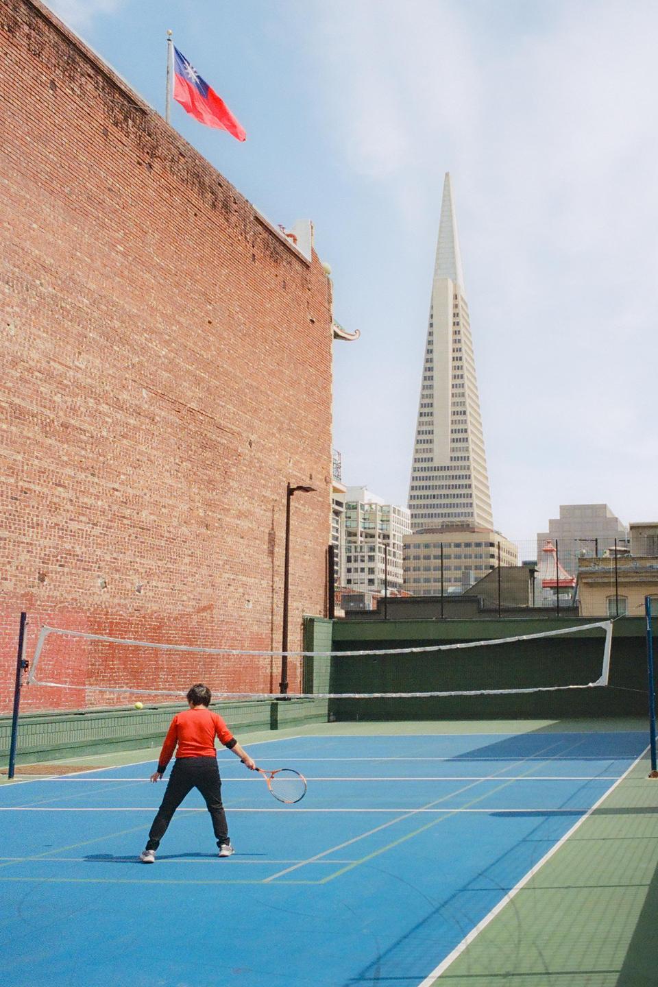 A person stands on a blue tennis court with a racket, facing a net, with a tall building and a flag visible in the background.