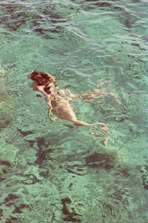 A woman swims in clear turquoise water, her body partially submerged and distorted by the water's surface.