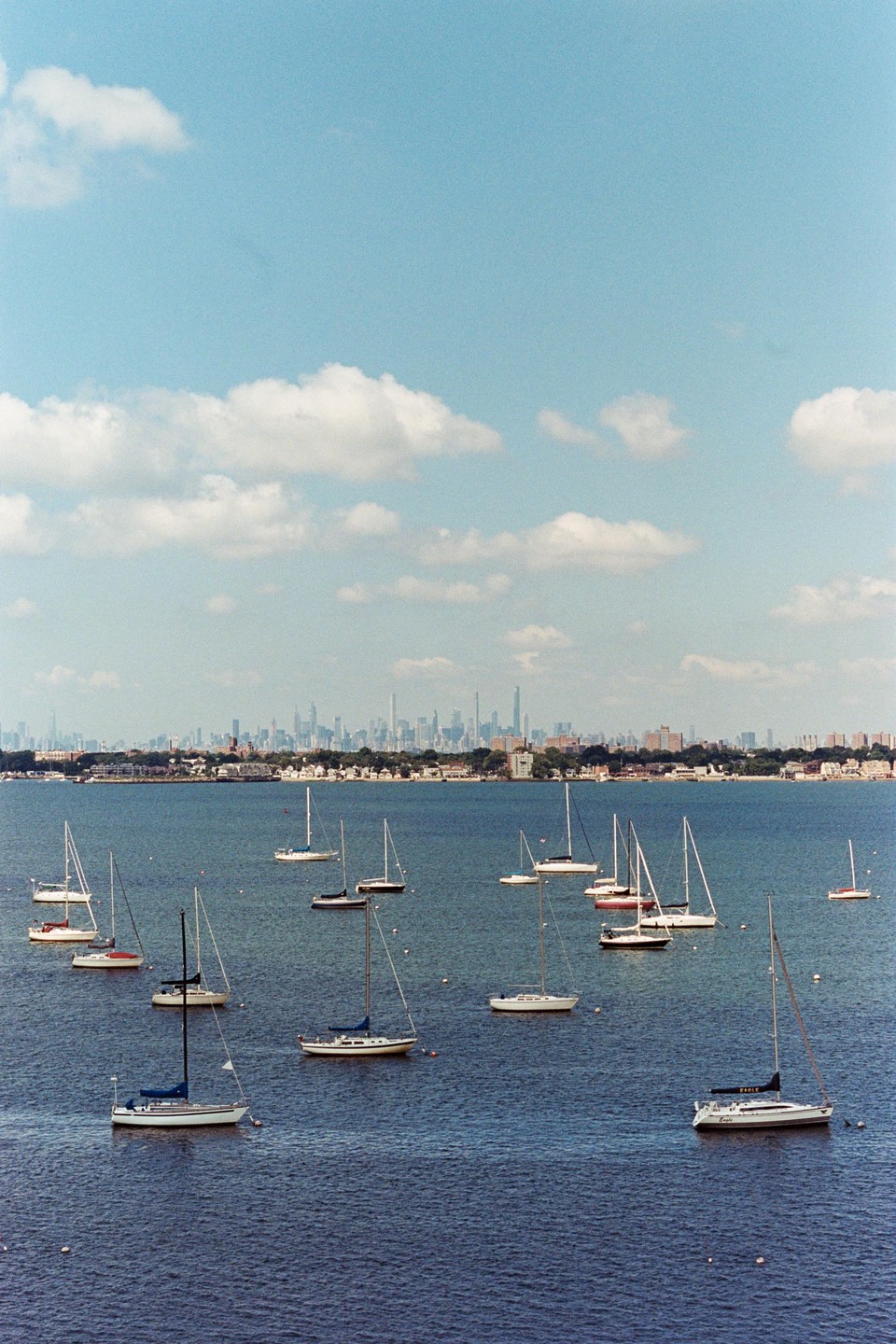 Sailboats are moored in a body of water with a distant city skyline under a partly cloudy sky.