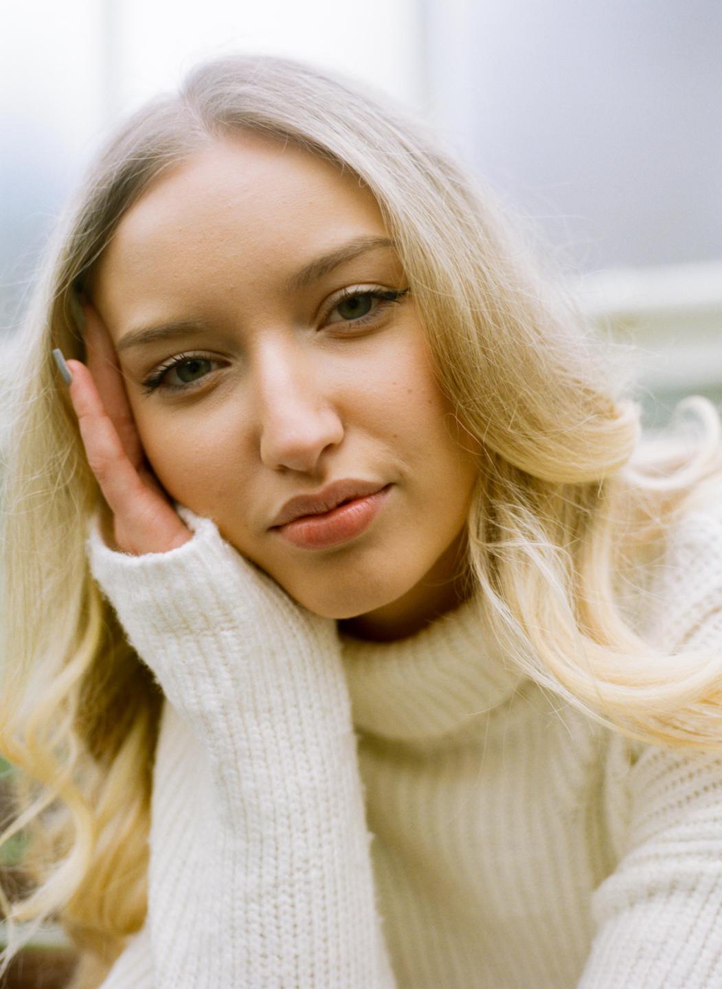 A woman with blonde hair and a white sweater rests her head on her hand.