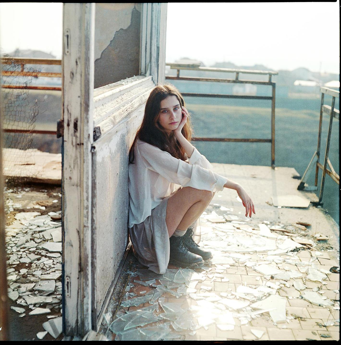 A woman crouches on a floor covered in broken glass, leaning against a weathered wall.