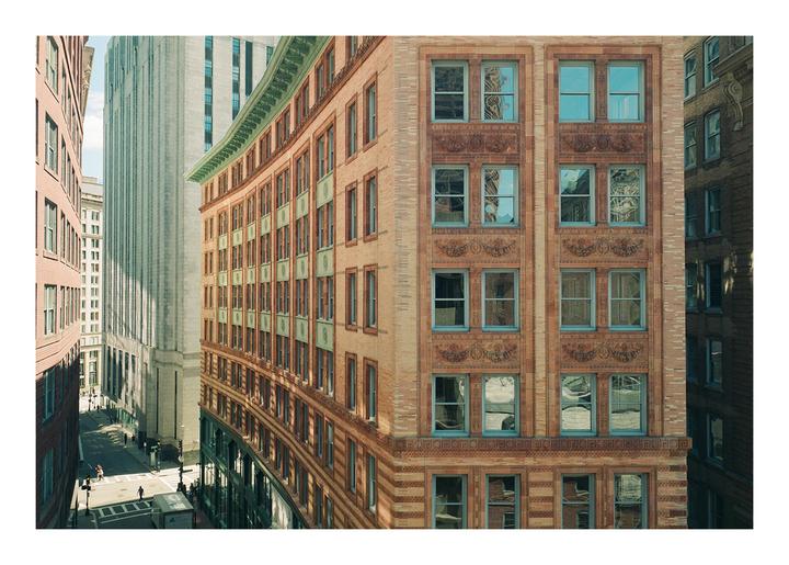 A curved brick building with many windows stands next to a tall, light-colored building, with a street and other buildings visible below.