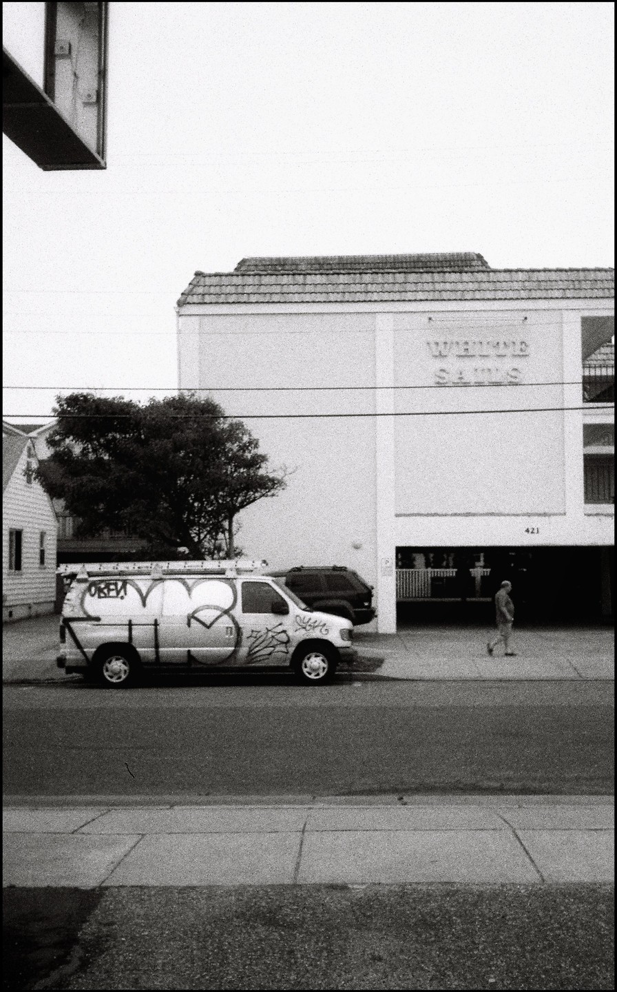 A man walks past a white van with graffiti parked on a street next to a building with "White Sands" written on it.