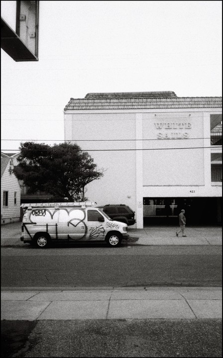 A man walks past a white van with graffiti parked on a street next to a building with "White Sands" written on it.