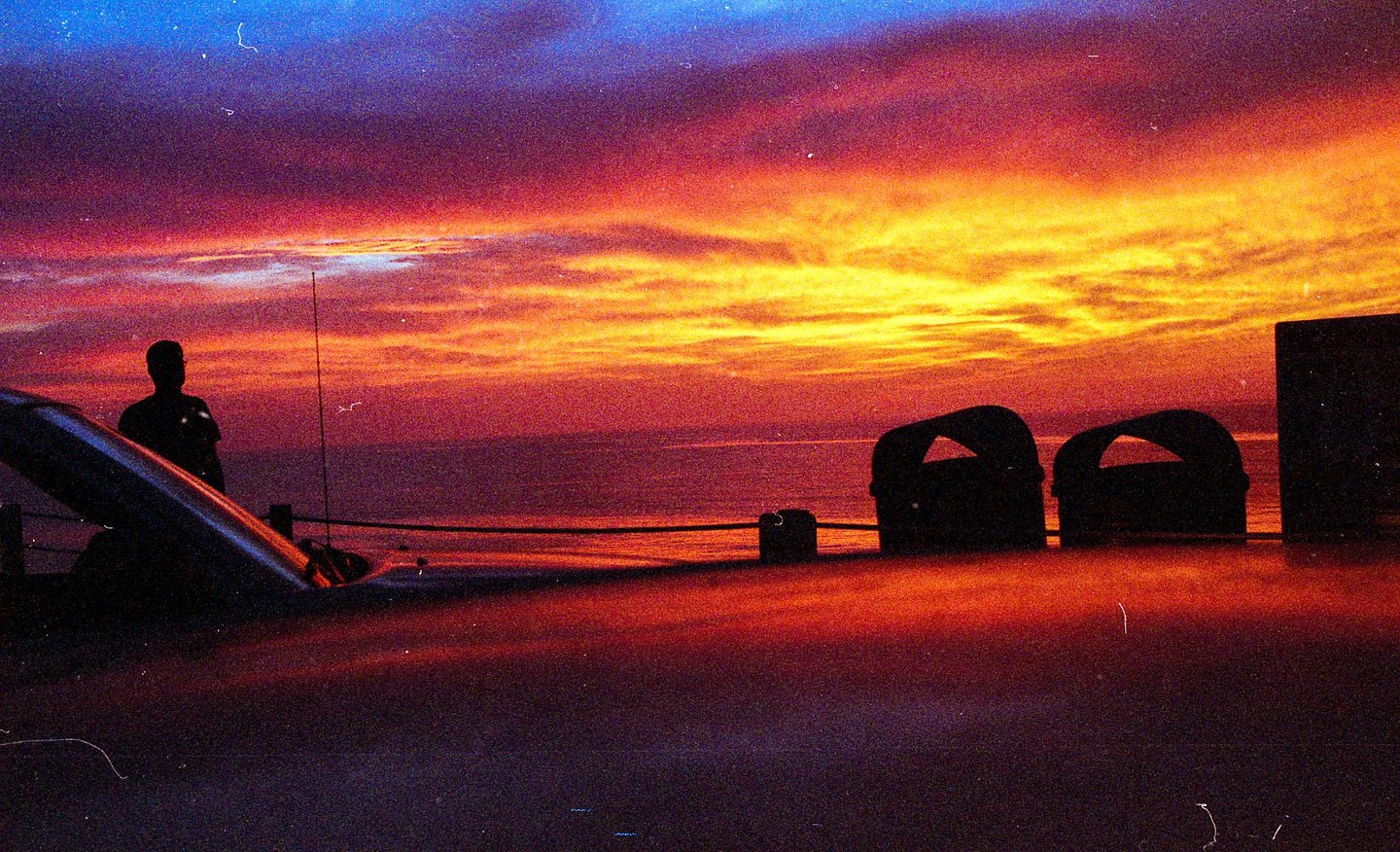 A man stands on a boat deck looking out at the ocean during a colorful sunset.