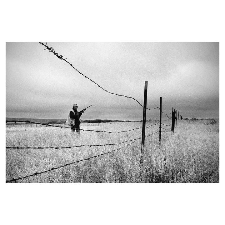 A man with a rifle stands in a field behind a barbed wire fence.