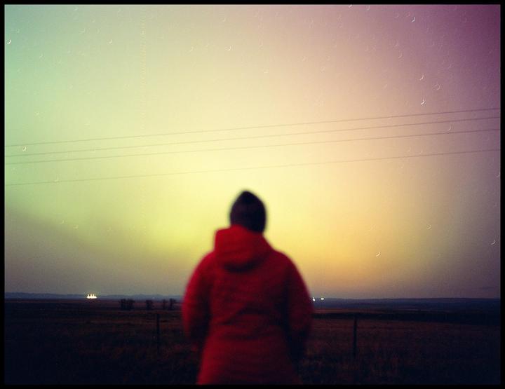 A person in a red jacket stands with their back to the camera, looking out at a colorful sky over a field.