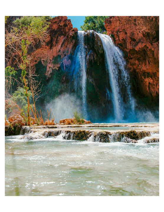 A large waterfall cascades over red rock formations into a series of pools and a river.