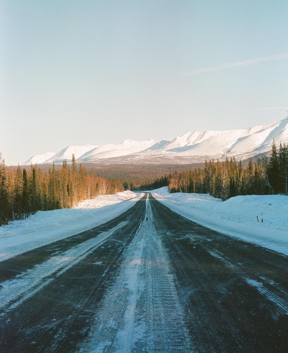 A long, snow-covered road stretches into the distance, flanked by evergreen trees and snow-capped mountains under a clear sky.