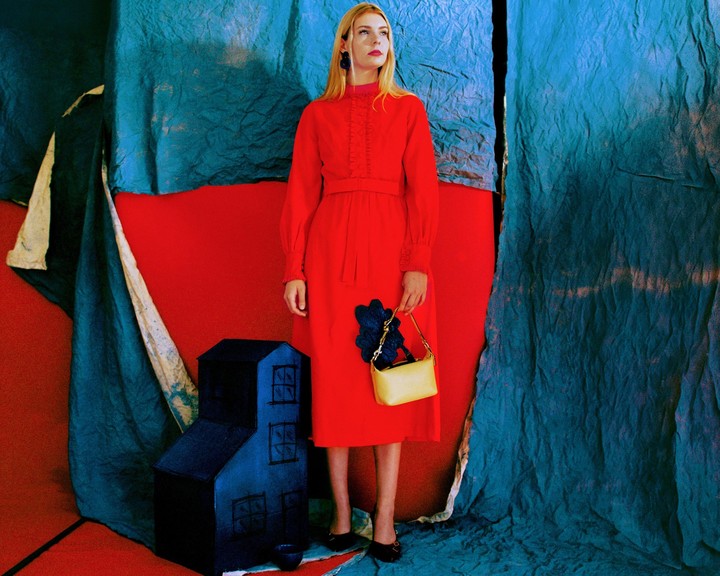 A woman in a red dress holds a yellow handbag while standing next to a blue house prop against a red and blue backdrop.