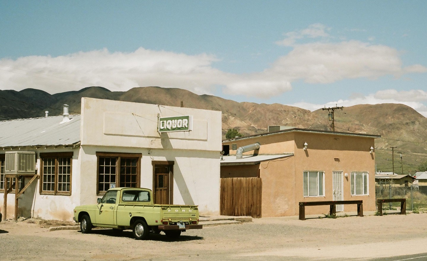 A light green pickup truck is parked in front of a liquor store with mountains in the background.