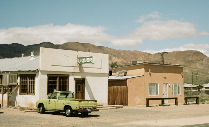 A light green pickup truck is parked in front of a liquor store with mountains in the background.