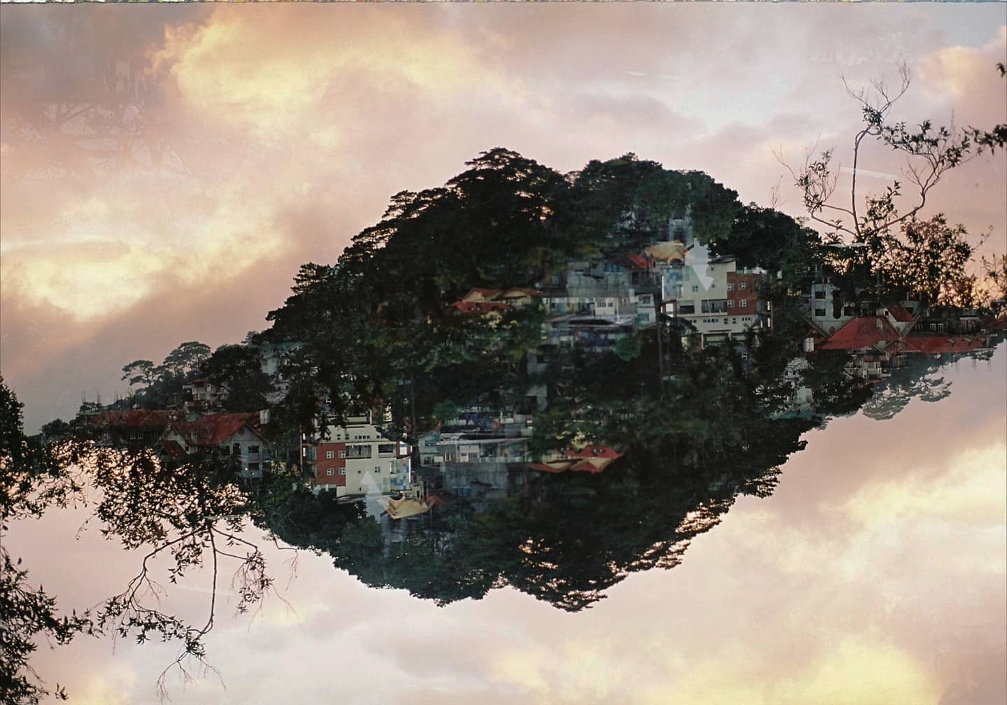 A double exposure shows a mountain covered in trees and buildings floating in a cloudy sky.