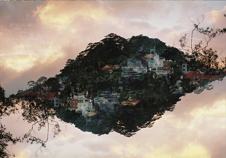 A double exposure shows a mountain covered in trees and buildings floating in a cloudy sky.