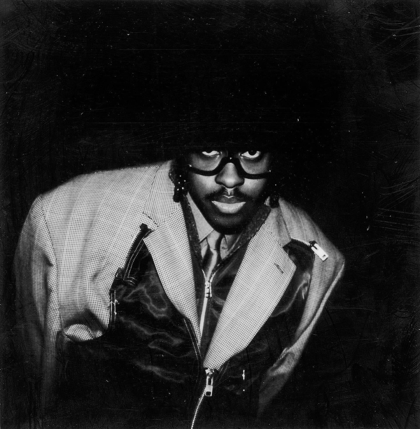 A man with glasses and a large afro wears a patterned jacket over a zippered vest and tie.