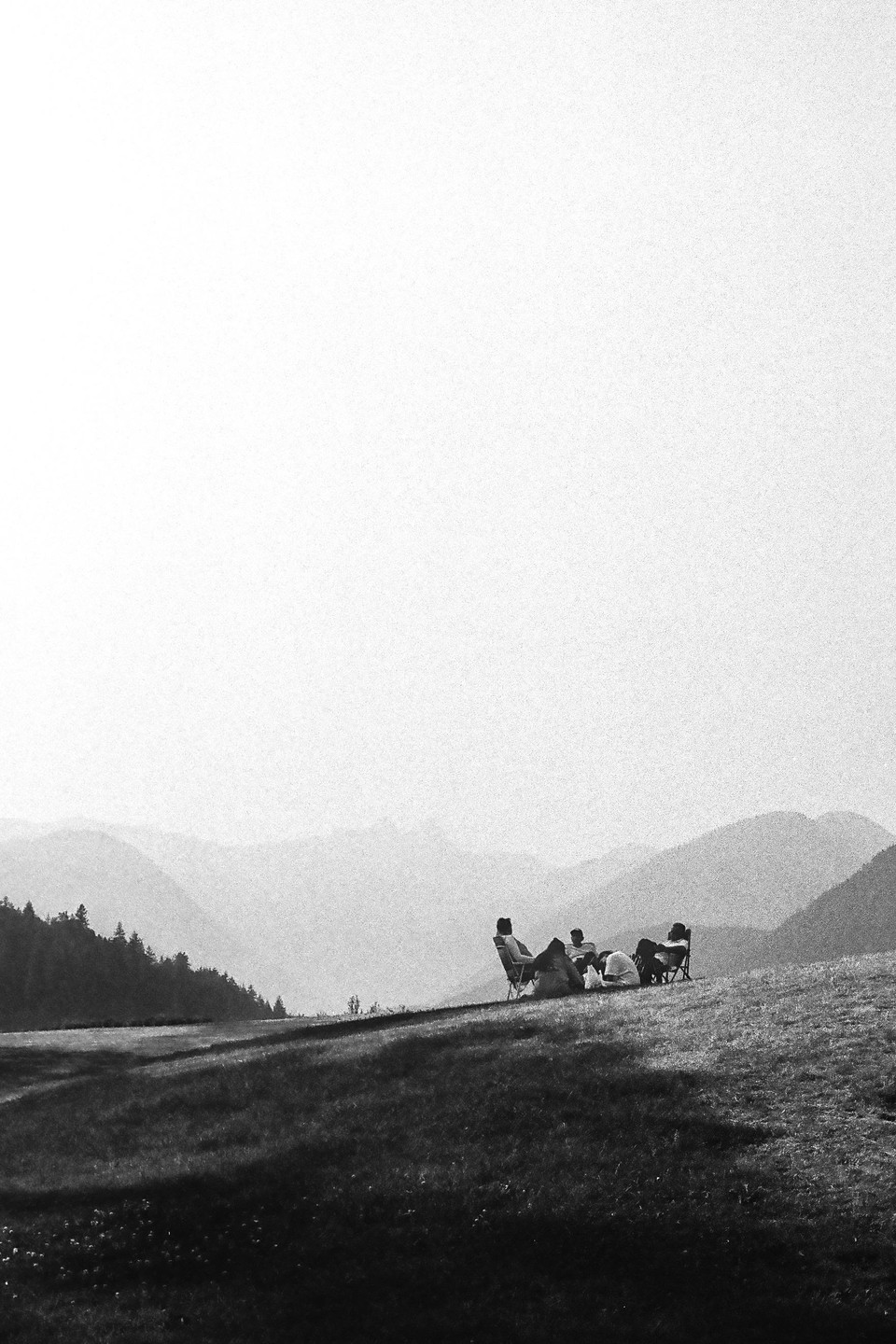 Four people are sitting in chairs on a grassy hill with mountains in the background.